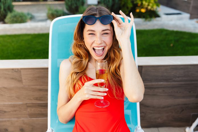 Smiling young woman lies on deck chair drinking champagne.
