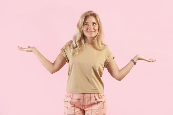 Caucasian blonde woman smiles and spreads her hands gesturing no idea isolated on pink background.