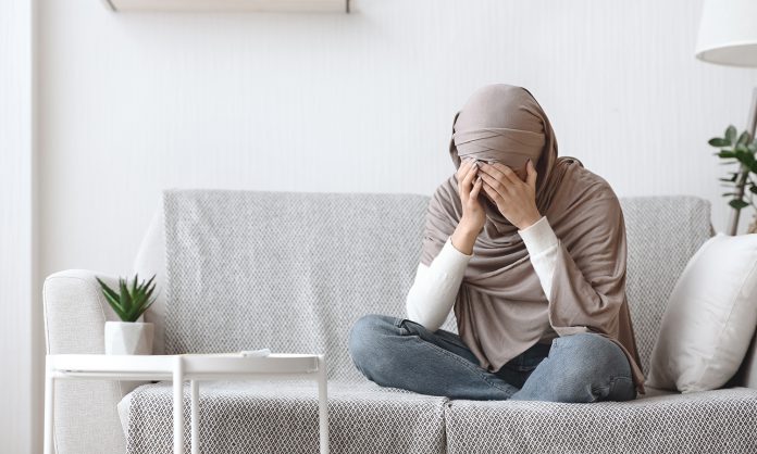 Upset muslim woman in headscarf crying on couch at home
